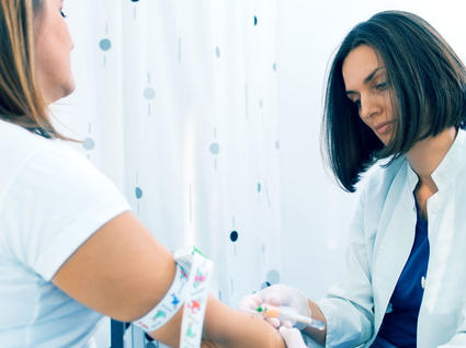 Healthcare professional in white coat drawing blood from a patient's arm. The patient wears a white shirt and has a colorful tourniquet around their upper arm