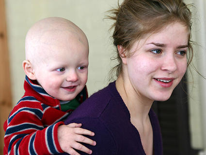 Child with a bald head, is smiling while playfully holding onto the woman’s shoulders. The woman looks content as she interacts with the child.