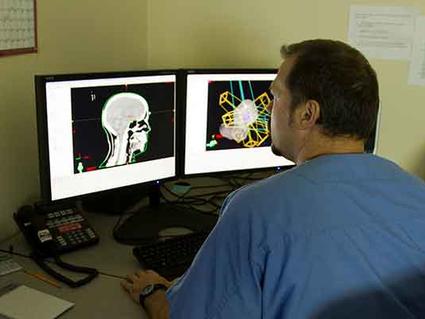 Medical professional in scrubs reviewing brain scan images on dual computer monitors, showing both traditional X-ray and enhanced colorized views.