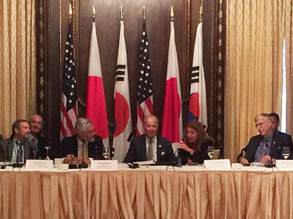 Political and health leaders sit at table with the flags of USA, Japan, and Korea behind them