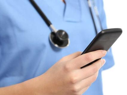 A close-up of hand holding a cell phone. The person’s body, wearing blue scrubs and a stethoscope around their neck, is blurry yet visible in the background.