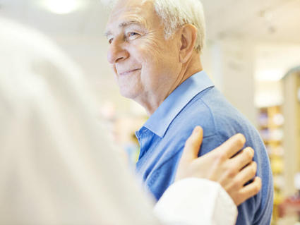 Smiling elderly man in blue shirt receiving a supportive hand on his shoulder from someone off-camera 