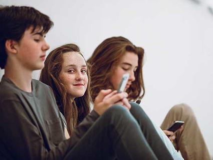 Three teenagers sitting together, with the girl in the middle smiling at the camera while her friends look at their phones in a casual, relaxed setting.