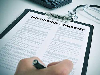 A close-up of a hand holding a pen to a clipboard holding a piece of paper. The words “Informed Consent”  are printed in large letters at the top of the paper, with smaller text in two columns below. A stethoscope and part of a keyboard are in the background.