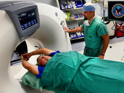 Patient lying on CT scan table receiving low-dose screening for lung cancer, with medical technician operating the machine in a hospital setting displaying Veterans Affairs and ICARE logos on equipment monitors.