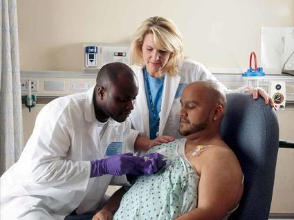 Two medical professionals providing chemotherapy treatment to a patient who has a port in his chest, with one healthcare worker administering the treatment while the other provides support in a hospital setting.
