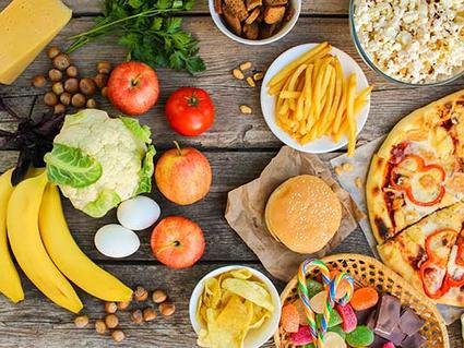 Overhead view of various foods arranged on wooden surface, including fresh produce like bananas, apples, and cauliflower on the left side, and processed foods like pizza, burgers, french fries, and snacks on the right side.