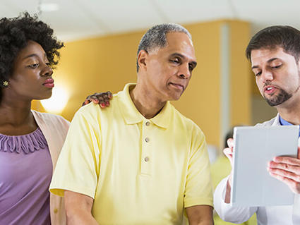Healthcare provider in white coat explaining information on a tablet to a man in yellow shirt and woman in purple blouse in a medical setting.