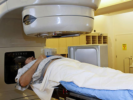 A patient lays on his back under a radiation machine. The patient is dressed in a hospital gown and covered with a blanket. The machine is large and grey with a cylindrical section directly above the patient’s chest. The background is tinted yellow and there are cabinets and a door.