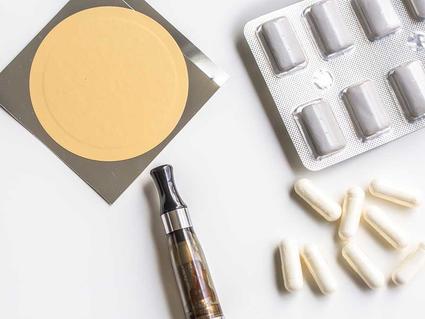 Various smoking cessation products including an e-cigarette device, nicotine patches, pills in blister packs, and white capsules arranged on a white surface.