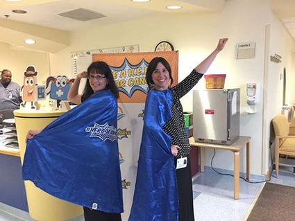 Becky Owens (left) and Sara Rothschild (right) smiling and wearing bright blue superhero capes pose in a hospital environment. The capes feature the words "Be a Hero for Cancer."