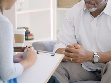 Seated female health care provider with clipboard meets with seated male patient.