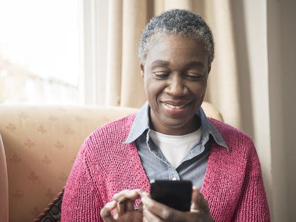 Woman with short hair sitting comfortably in a chair by a window, smiling while using a smartphone. She is wearing a pink cardigan over a denim shirt. 