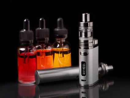 An image of an e-cigarette machine in the foreground with three small bottles that have eyedropper tops in the background. The small bottles contain liquids colored red, orange and yellow; the e-cigarette machine is grey with metal touches.