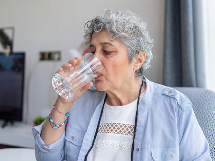 A person with short gray hair drinks water from a tall glass.