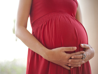 A close-up of a woman’s arms holding her pregnant belly. She is wearing a red dress.