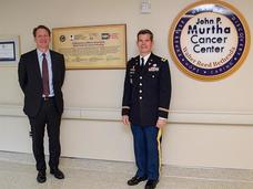 NCI Director Norman Sharpless, M.D. (left) standing with Colonel Craig Shriver, M.D.  in a hallway next to a sign for the John P. Murtha Cancer Center. The wall displays a circular emblem and a plaque with text about the center.