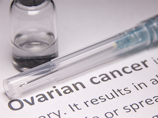 A close-up of a syringe and medical bottle containing clear liquid. These two items are sitting on a piece of paper that shows the definition of ovarian cancer. The words “ovarian cancer” are bold; the rest of the text is mostly cropped off the image, though what is shown indicates it is a definition of ovarian cancer.