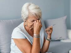 An elderly woman sits on a couch holding her glasses in one hand and rubbing her forehead with the other, appearing to be in discomfort.