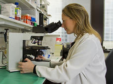A female scientist sits down and looks through a microscope. She wears a lab coat and has shoulder length dirty blonde hair. She has a happy expression on her face. In the background, shelves with various types of equipment and binders can be seen, as well as large windows with the blinds pulled down, indicating a lab environment.