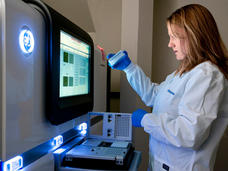 A female research technician holds up a small instrument in front of a large machine at the Advanced Technology Research Facility at NCI's Frederick National Laboratory for Cancer Research. The technician is wearing a white lab coat and blue gloves and has shoulder length brown hair with highlights. The large machine has a screen and lights on it; the instrument being held fits in to a smaller device that is below the screen.