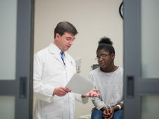 A doctor looks at a piece of paper and speaks to his patient. The doctor is a white middle-aged male, wearing a lab coat, button up shirt and tie, He is standing and gesturing as he speaks. The patient, a younger Black woman with glasses, a long sleeve grey sweater and jeans, sits on the exam table and looks slightly distressed. Out of focus doors to the room are in the foreground.