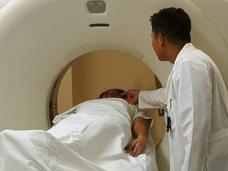 A doctor in a white coat watches as a patient covered in a white blanket goes into a CT machine. The doctor’s hand is partially in the machine.
