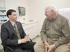 A doctor and a patient, both sitting, speak to each other. The doctor is middle aged with short dark hair, glasses, a grey suit and tan shirt with patterned tie. He’s sitting in a chair. The patient is an elderly man in a green button up shirt and khakis with white hair; he sits on an exam chair. A sink, cabinets, and gloves are in the background.