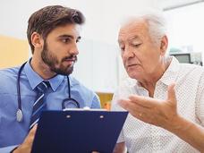 Doctor with stethoscope listening attentively to an elderly male patient who is speaking and gesturing with his hands during a consultation.