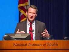 Ned Sharpless, dressed in a suite and tie, stands speaking at a podium. His expression is neutral and his hands are raised to chest level. The podium is wooden and has text reading “National Institutes of Health” carved into it. The podium also has two microphones. In the background, an American flag and a curtain can be seen.