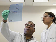 Two laboratory researchers examining a DNA gel or test results together under fluorescent lighting, both wearing protective eyewear and lab coats.