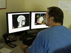 Medical professional in scrubs reviewing brain scan images on dual computer monitors, showing both traditional X-ray and enhanced colorized views.