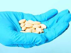 An outstretched hand wearing a blue latex glove holds a handful of oval pills which are light orange in color.