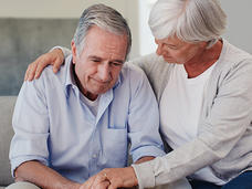 Elderly woman comforting a man by placing her hand on his shoulder as they sit together on a couch, with the man appearing distressed or contemplative.