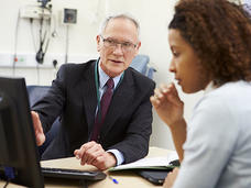 A doctor points at a computer screen while speaking to a patient. The doctor is an elderly man with white hair, glasses, and a dark suit with a lanyard around his neck. He is looking at the computer screen, not the patient. The patient is an adult woman with curly brown hair and a long light blue shirt.