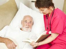 Nurse in pink scrubs reading to an elderly male patient in bed as part of palliative care, focusing on comfort and quality of life during cancer treatment.