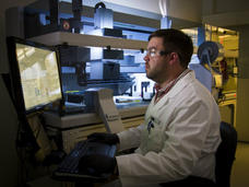 A scientist in a lab coat and safety glasses is working at a computer station in a laboratory. The lab is equipped with advanced machinery in the background. The scientist is focused on data displayed on the screen.
