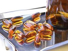 Golden-brown gel capsules sit on a metal surface. Part of a brown, glass pill bottle sits near the capsules.