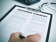A close-up of a hand holding a pen to a clipboard holding a piece of paper. The words “Informed Consent”  are printed in large letters at the top of the paper, with smaller text in two columns below. A stethoscope and part of a keyboard are in the background.
