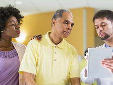 Healthcare provider in white coat explaining information on a tablet to a man in yellow shirt and woman in purple blouse in a medical setting.