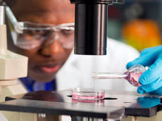 A male researcher, wearing lab goggles, a white coat, and blue gloves, pours pink fluid from a small glass container into another small cylinder that is focused underneath a microscope. The microscope and fluid containers are in focus in the foreground, the man and his environment are blurry, indicating depth of field.