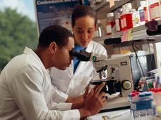A male researcher looking through a microscope in a laboratory with a female researcher standing near.