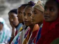A group of individuals is sitting in a line, with one person looking directly at the camera. They are wearing colorful clothing and head coverings, appearing thoughtful.