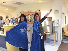 Becky Owens (left) and Sara Rothschild (right) smiling and wearing bright blue superhero capes pose in a hospital environment. The capes feature the words "Be a Hero for Cancer."