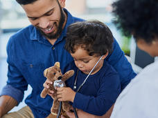 A pediatric patient wears a stethoscope and holds a stuffed bear as his father smiles at him