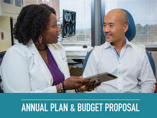A doctor speaks to a patient in her office. The doctor is a Black woman with shoulder length hair, a white coat, a purple blouse, and jewelry. She is holding a tablet. The patient is a bald Asian man wearing a white button up shirt. Both are sitting and wear happy expressions. In the background, a scan can be seen on the computer screen, along with windows and cabinets. Text at the bottom reads “Annual Plan & Budget Proposal”.