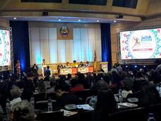 A panel session being held in a small auditorium. There are five panelists, seated, and one moderator, standing at a podium. Large screens on either side of the stage have the branding of the conference. The backs of the heads of the audience can be seen int he foreground, and they are darker than the stage.