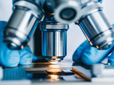 Close-up of a microscope being operated by a person wearing blue gloves. The microscope's lens is focused on a glass slide with light illuminating the sample.