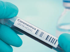A close-up of glove-covered fingers holding a sample vial with the label “Coronavirus (2019-nCoV)” along with a bar code. The vial has a very dark liquid. In the background, out of focus, is a clear bottle with blue top.