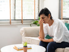 A picture of an older woman sitting on a couch, with one hand on her temple. 